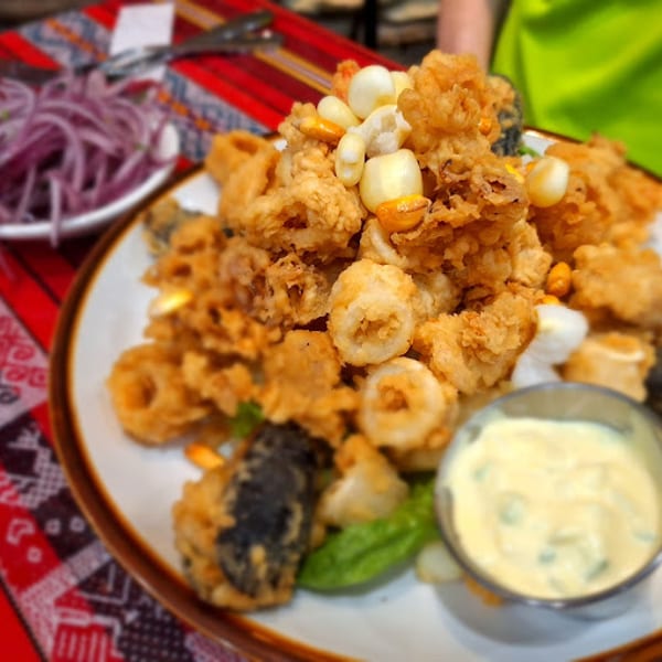 Plate of golden fried calamari and seafood with corn, dipping sauce, and red onions on a patterned tablecloth.