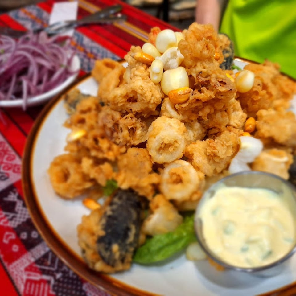 Plate of golden fried calamari and seafood with corn, dipping sauce, and red onions on a patterned tablecloth.